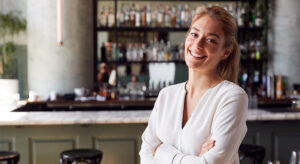 Mulher branca e loira sorrindo em um restaurante com os braços cruzados.