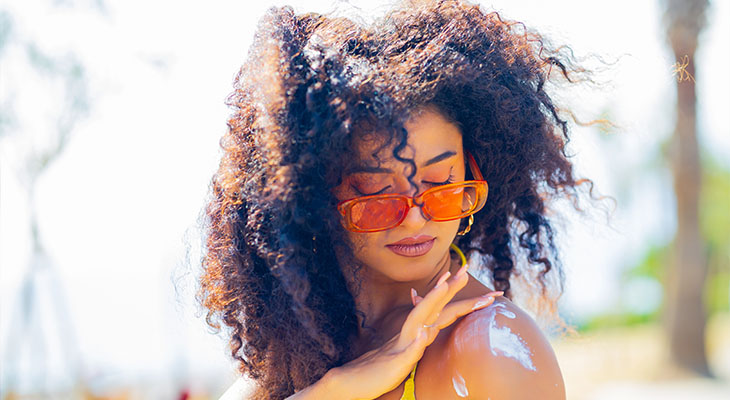 Mulher na praia, com óculos de sol, passando protetor solar em um dos ombros.